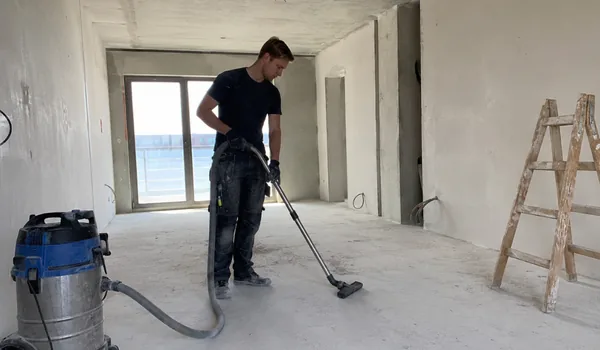 Ehitustolmu eemaldamine betoonpõrandalt tööstusliku tolmuimejaga, lõpuringil tõmbab koristaja valge puuvillase kindaga üle aknalaudade ja uksepiitade ning iga tolmujäljega pind puhastatakse kohe uuesti.