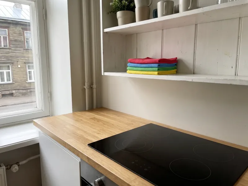 Kalamaja puumaja korteri köök pärast iganädalast hoolduskoristust, tammepuidust tööpind ja must klaaskeraamiline pliit, lahtisel riiulil kokkuvolditud punane, sinine, roheline ja kollane mikrokiudlapp, aknast paistab vana puumaja, kõrbenud jäägid kaabitakse pliidiplaadilt maha pliidikaabitsaga enne niisket pühkimist.