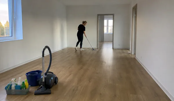 Tühja korteri koristus, koristaja peseb põrandat mopiga, esiplaanil tolmuimeja ja vahendid, väljalülitatud külmiku ukse jätab koristaja tühjas korteris paokile, et suletud külmikusse ei tekiks hallitust.